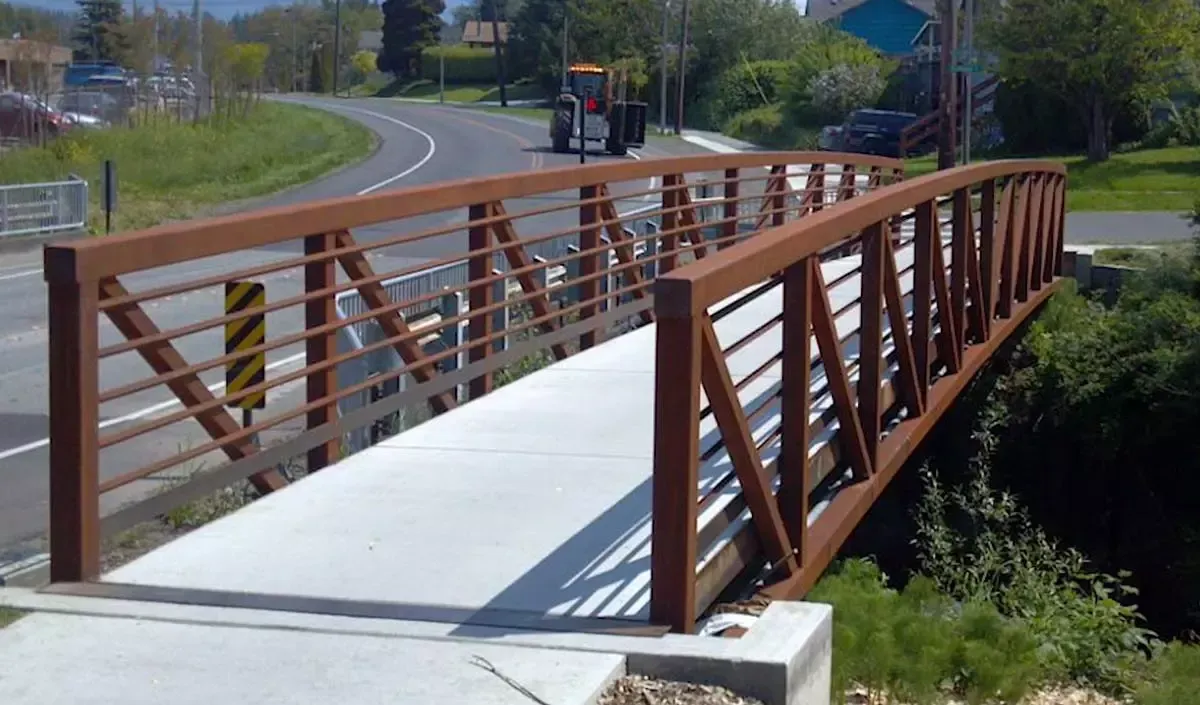 Modern pedestrian bridge with weathered steel railings and a concrete deck crossing a small ravine alongside a suburban road, with trees and houses in the background.