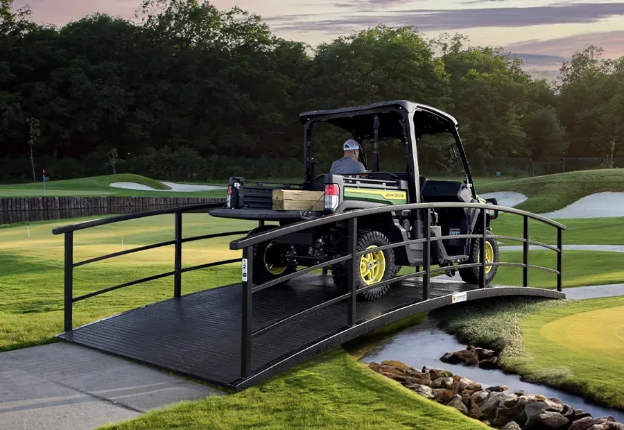 Curved modular cart bridge with black steel rails and composite deck, carrying a utility vehicle on a golf course.