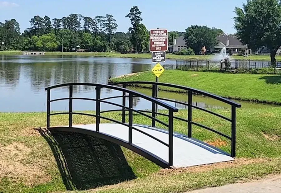 Prefabricated arched footbridge with powder-coated rails spanning a small swale near a pond fountain.
