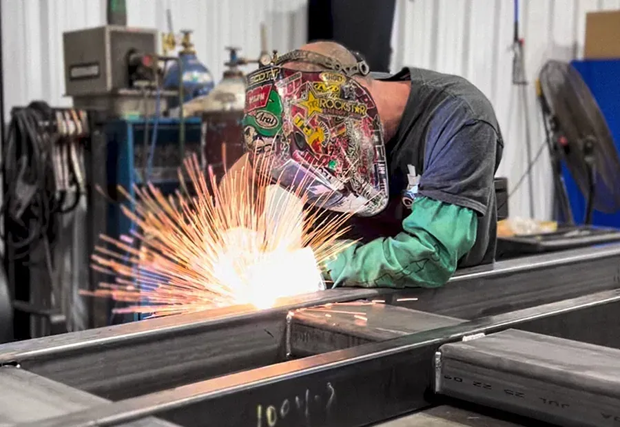 Welder joins structural steel for a modular bridge frame, sparks flying in the shop.