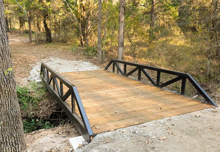 Compact trail bridge with triangular steel side trusses and stained wood deck in wooded setting.