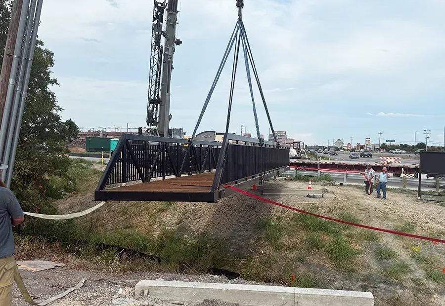 Crane sets a modular steel-truss pedestrian bridge with wood deck onto abutments by the roadway.