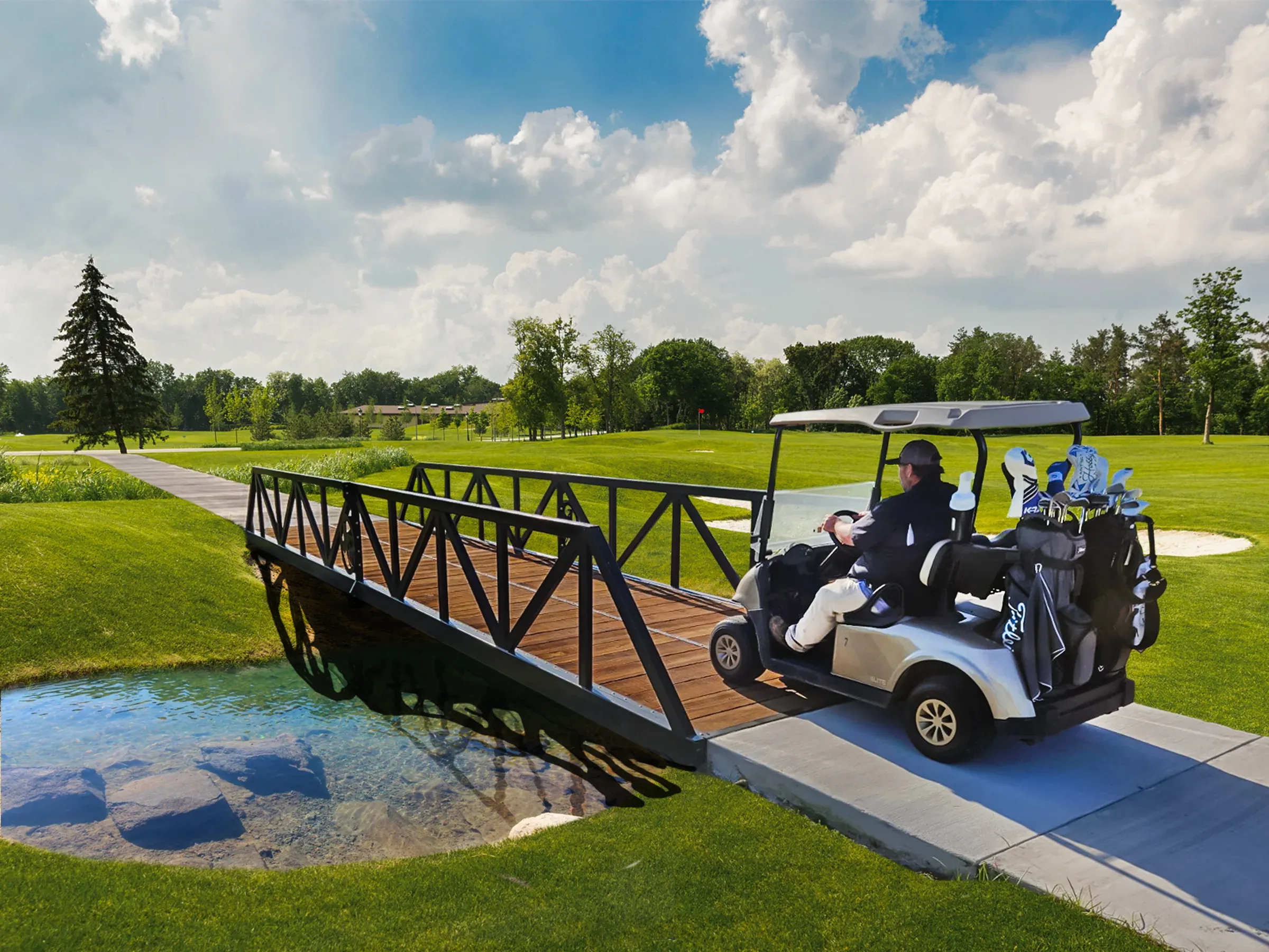 Man driving a golf cart across a steel-framed, wooden-deck bridge over a clear stream on a manicured golf course, surrounded by green fairways and trees under a partly cloudy sky.