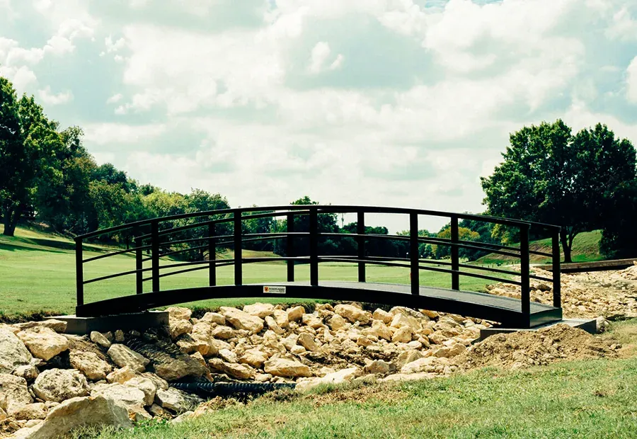 Curved modular cart bridge with black steel rails over a rock-lined drainage on a golf course.