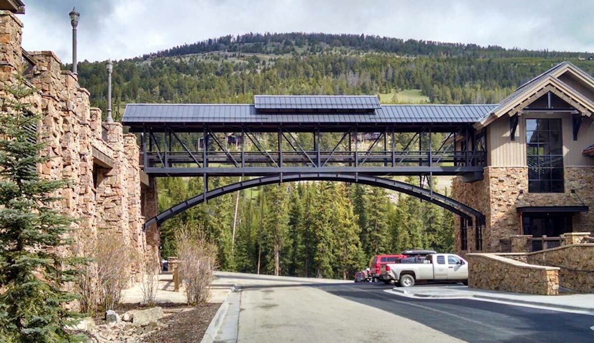 Modular covered walkway bridge with black steel trusses, arched underframe, alpine setting.