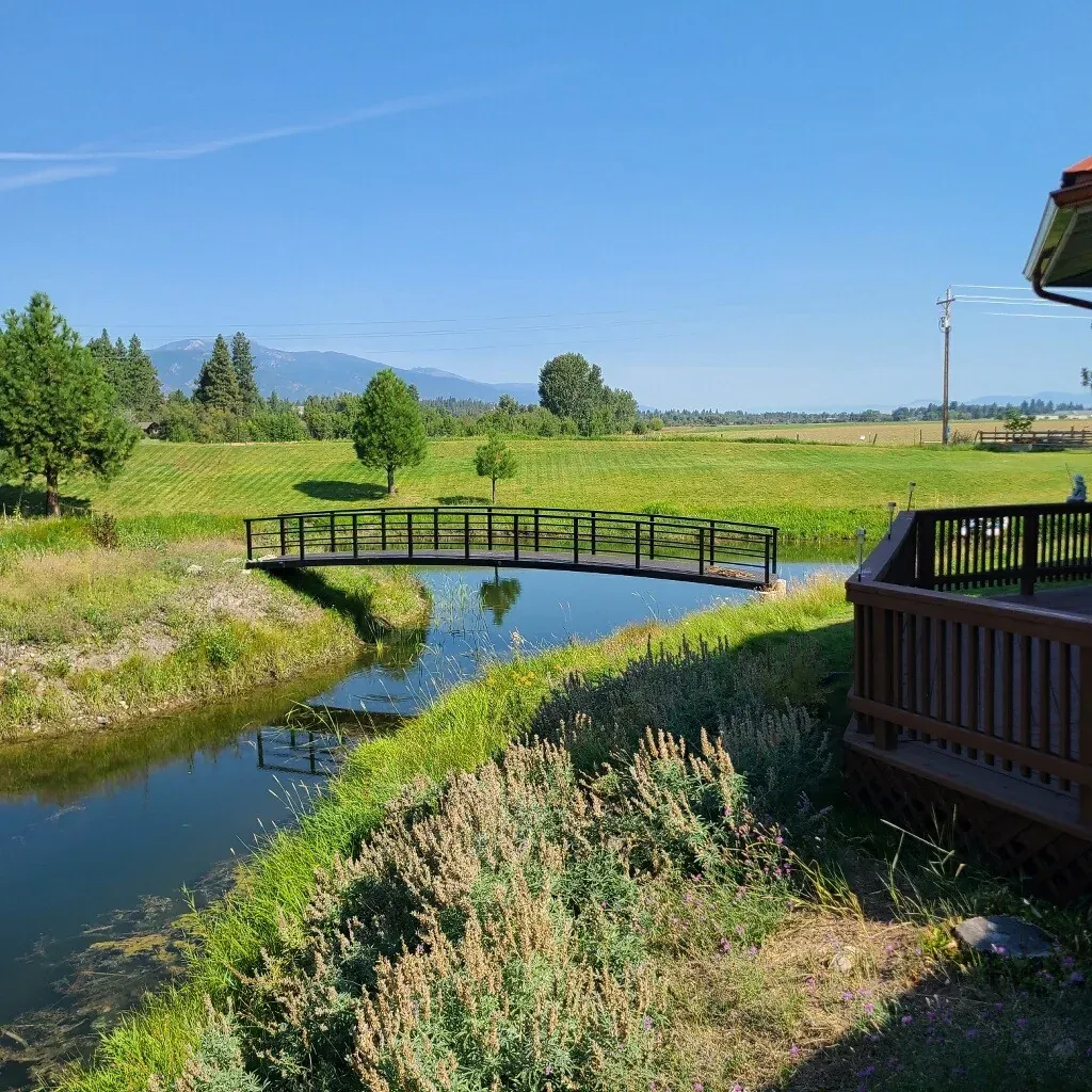 Curved pedestrian bridge with metal railings spanning a calm irrigation canal, set in a rural landscape with green fields, trees, and distant mountains under a clear blue sky. A wooden deck is visible in the foreground.