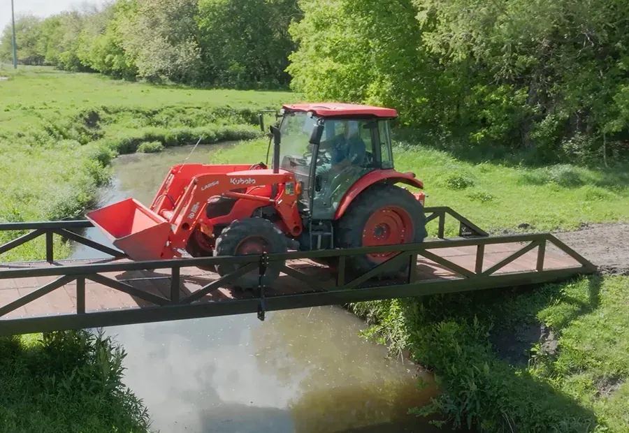 Modular steel-truss bridge with wood deck carrying a compact tractor over a creek.
