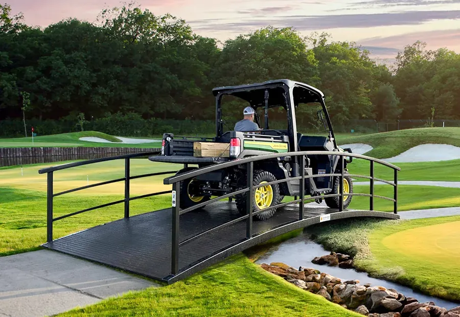 Curved modular cart bridge with steel rails and black deck, carrying a utility vehicle on a golf course.