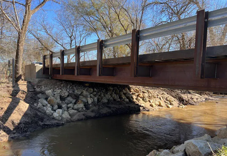 Modular steel beam span crossing a creek; stone slope protection at both ends.
