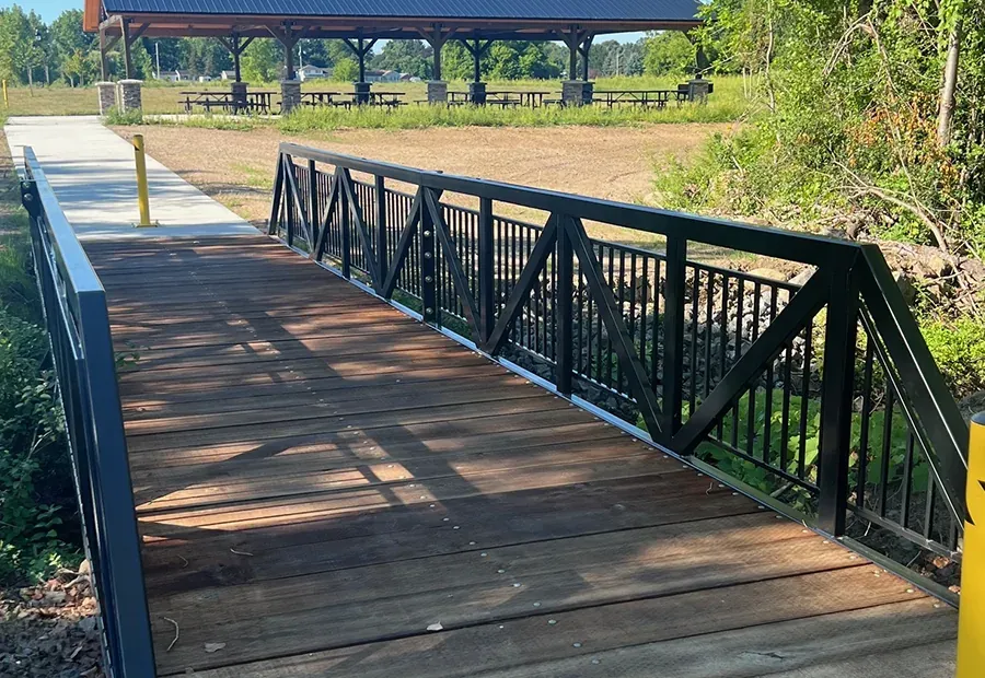 Modular steel-truss footbridge with wood deck connecting to a concrete path near a park pavilion.