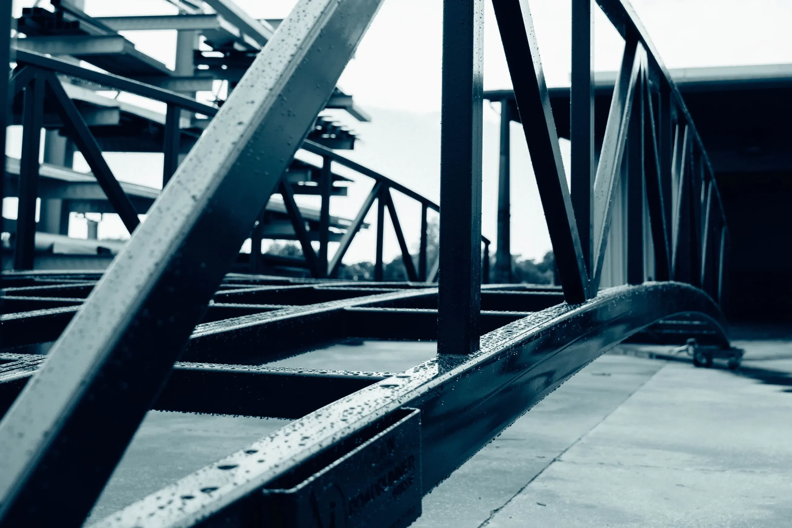Close-up view of a wet steel truss bridge frame in a fabrication yard, showcasing diagonal cross-bracing and structural details in a monochrome blue tone.