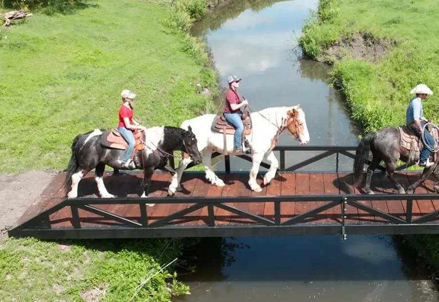 Modular steel-truss bridge with wood deck carrying horseback riders over a creek.